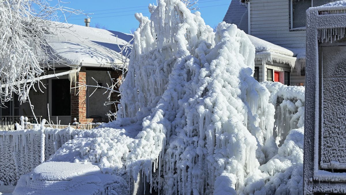 Residential street with cars, trees, and houses covered in thick ice and icicles on a clear winter day.