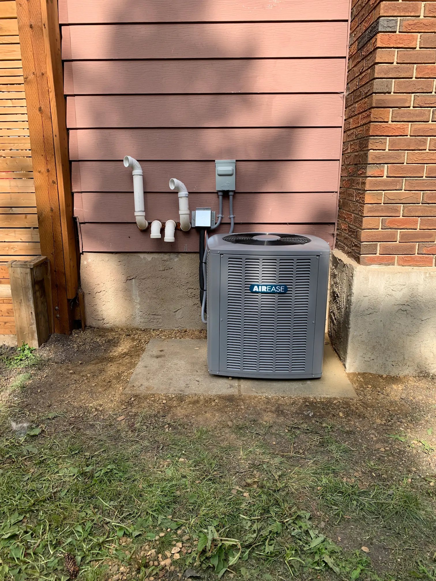 Outdoor air conditioning unit installed on a concrete pad next to a house with pink siding and a brick corner wall.