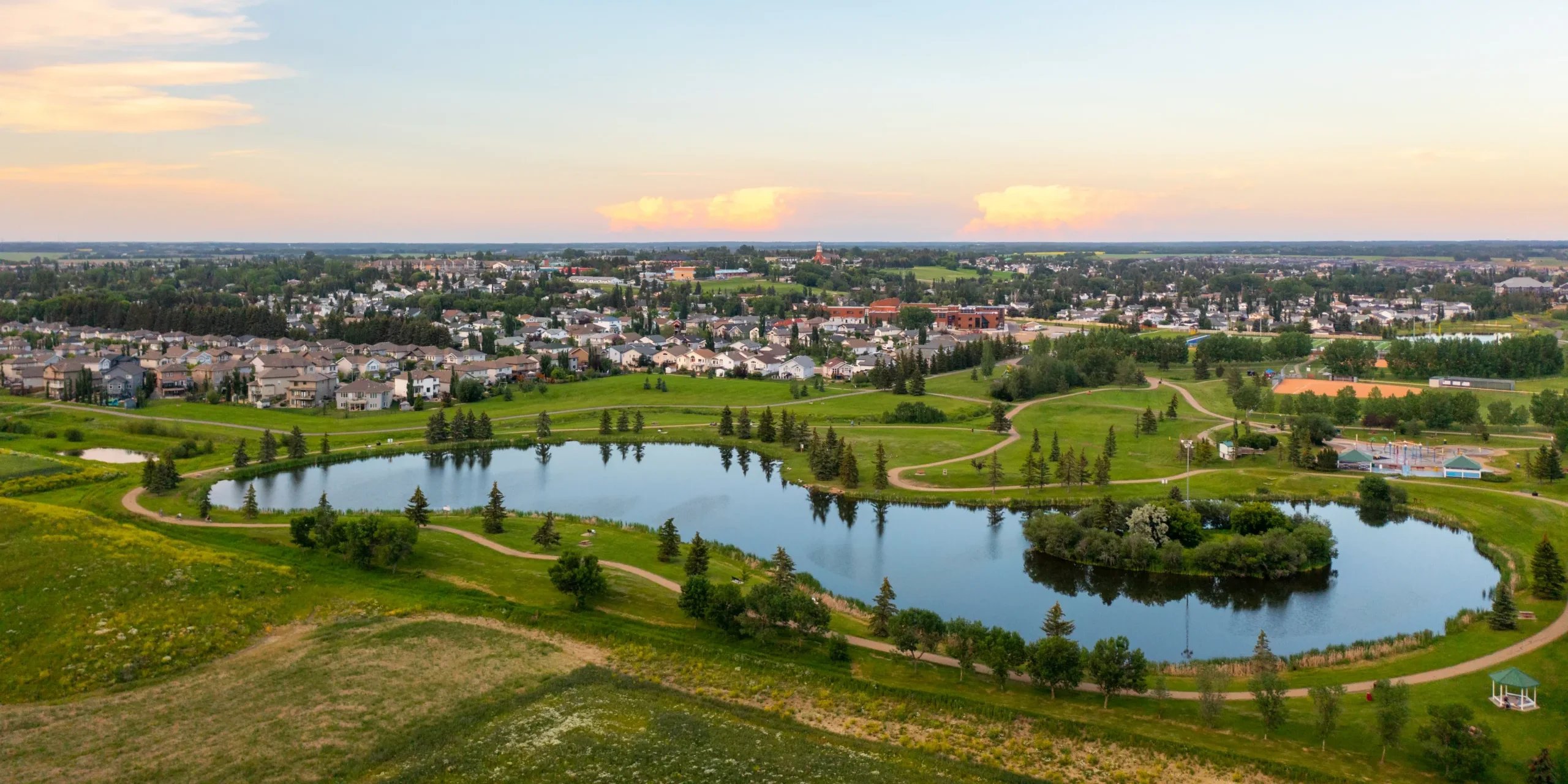 A suburban neighborhood near a park with a lake, walking paths, trees, and a playground at sunset.