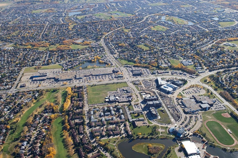 Aerial view of suburban area with houses, shopping centers, roads, a pond, and sports fields.
