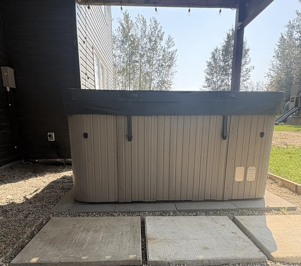 Backyard deck with glass railing, outdoor dining table with chairs, and storage space underneath containing stacked tires and firewood.