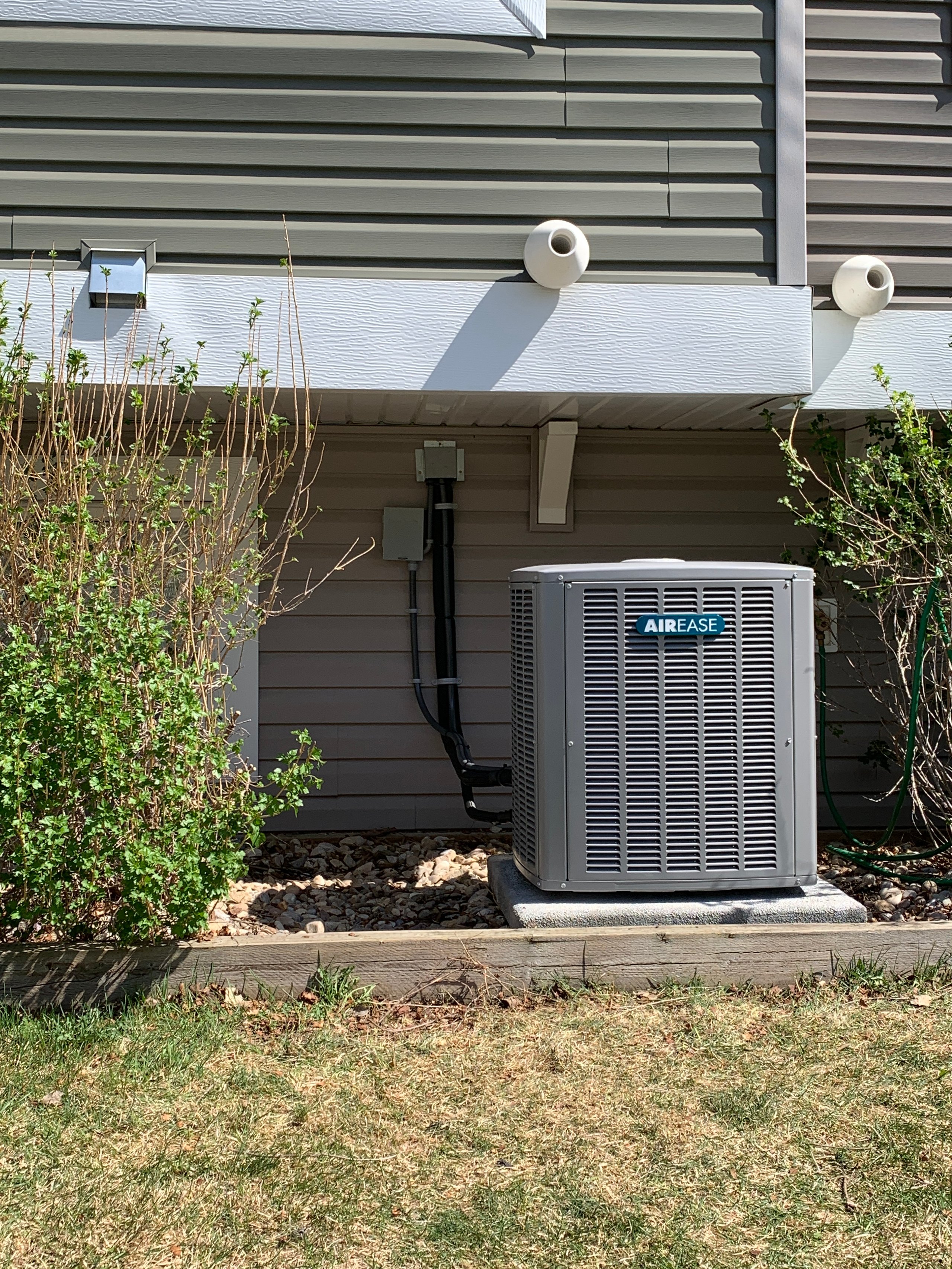 A gray Airease air conditioning unit installed on a concrete pad outside a house with beige siding and bushes on each side.