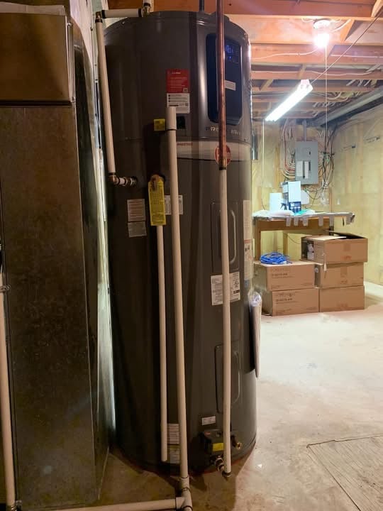 Large gray water heater with attached pipes in an unfinished basement with exposed ceiling beams and stacked cardboard boxes.