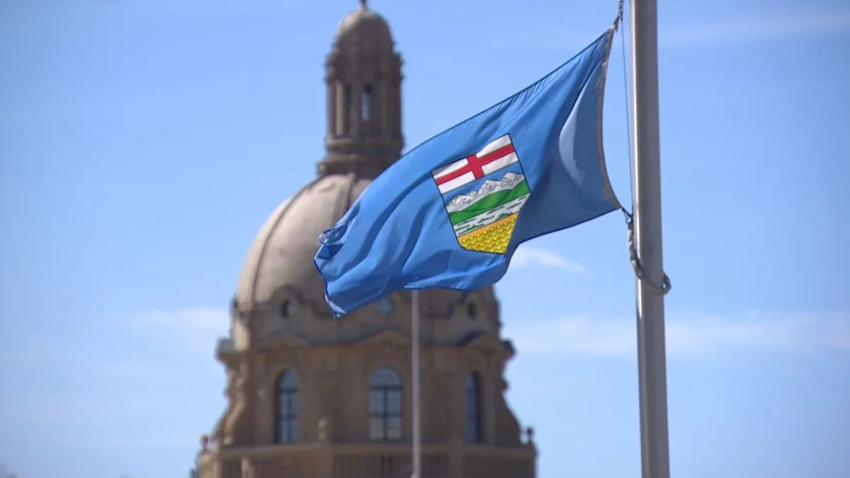 Blue flag with a shield emblem featuring red cross, mountains, green hills, and gold wheat blowing in front of a domed historic building under a clear sky.