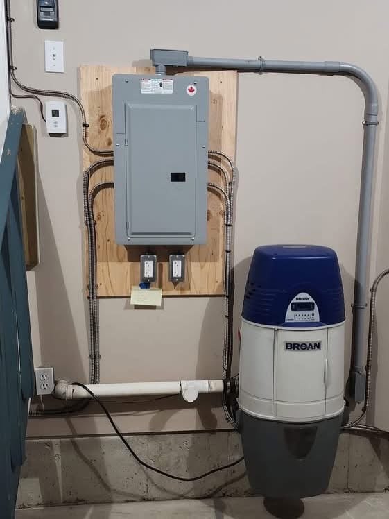 A residential electrical panel mounted on a wooden board with two outlets below it, next to a Broan central vacuum system with a blue and white body installed on a basement wall.