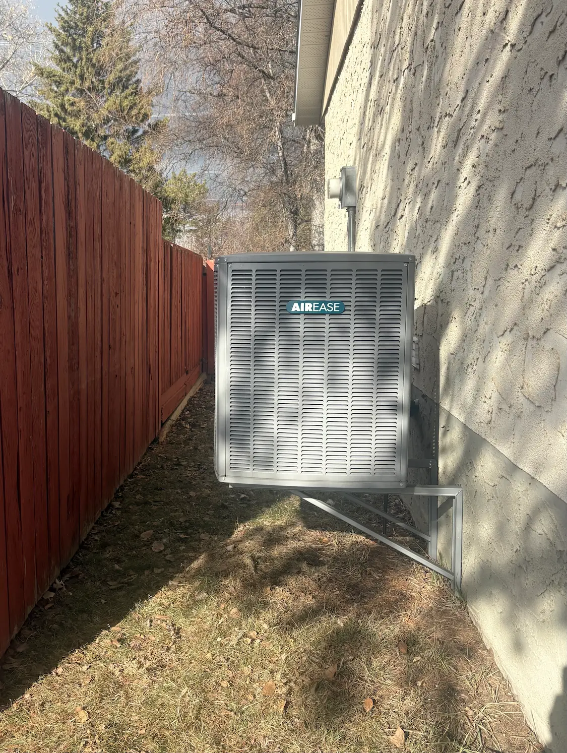 Outdoor AIR EASE air conditioning unit installed on a concrete slab against a beige house wall with connected electrical conduits.