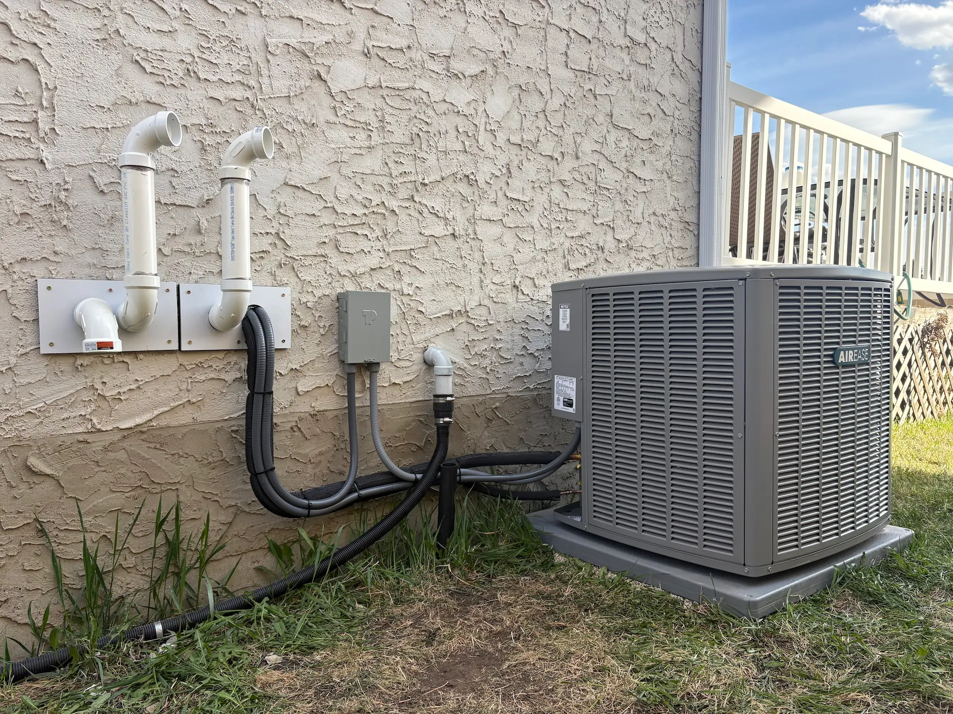 Two outdoor air conditioning units beside a wooden exterior wall on a concrete slab, next to green grass.