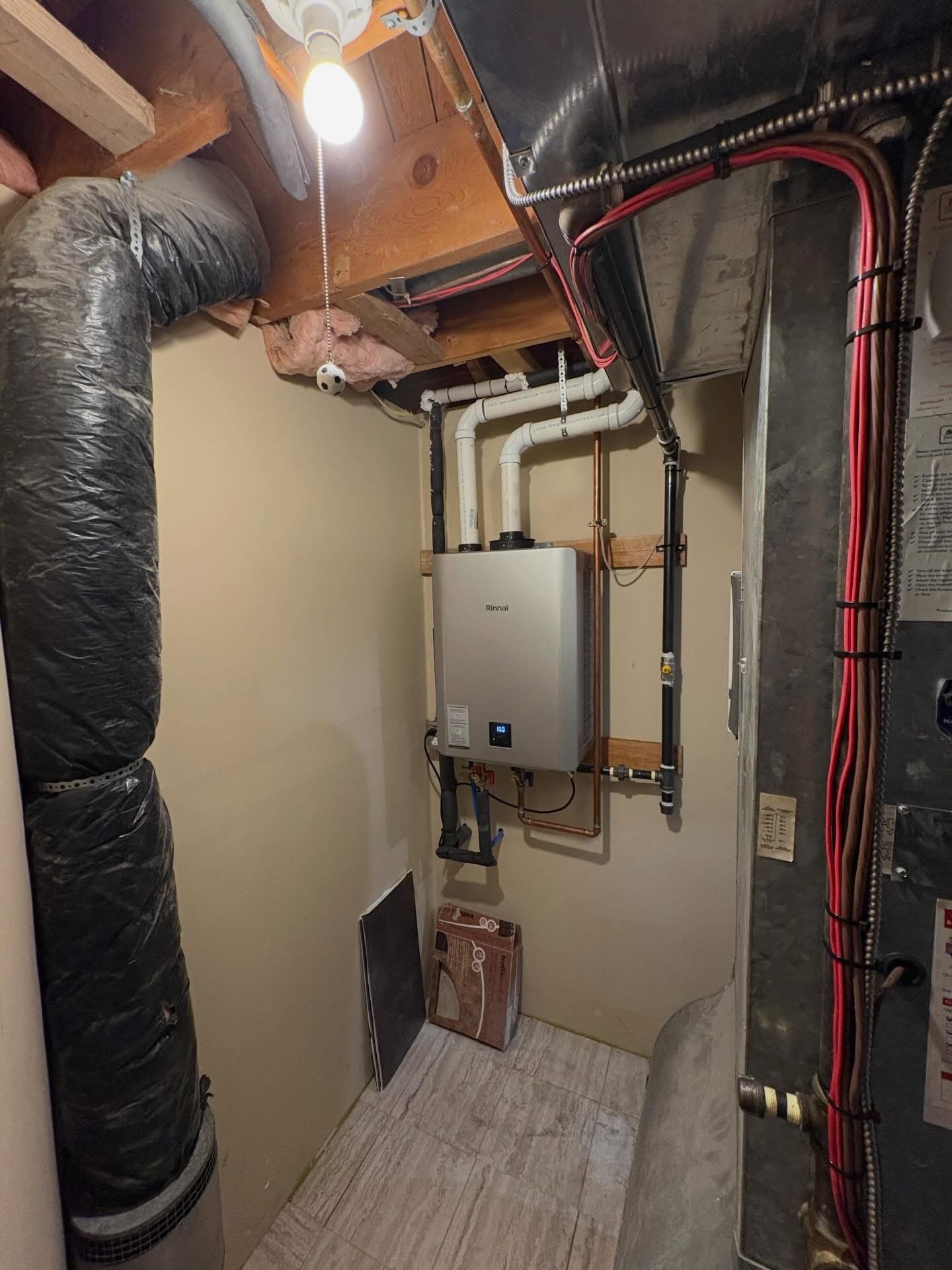 Small utility room with a Rinnai tankless water heater mounted on the wall, ductwork, pipes, and a light bulb hanging from the ceiling.