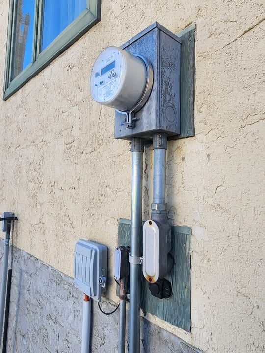 Electric meter and utility boxes mounted on the exterior wall of a beige stucco building under a window.