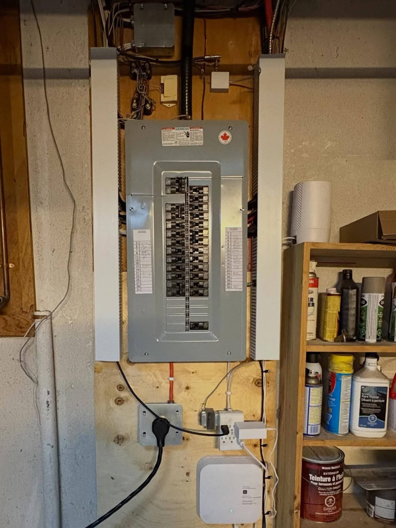Electrical breaker panel mounted on a wooden wall with various connected wires and outlets below, next to a shelving unit holding paint cans and sprays.