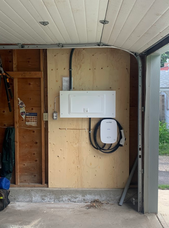 Electric vehicle charging station installed inside a garage on a plywood wall with coiled charging cable.
