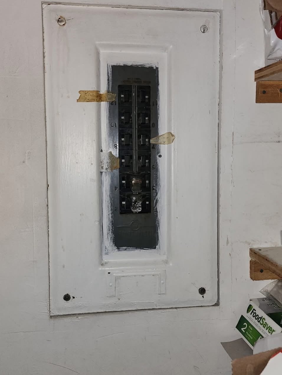 Old white electrical breaker panel mounted on a wall with several black switches and yellowed tape labels.
