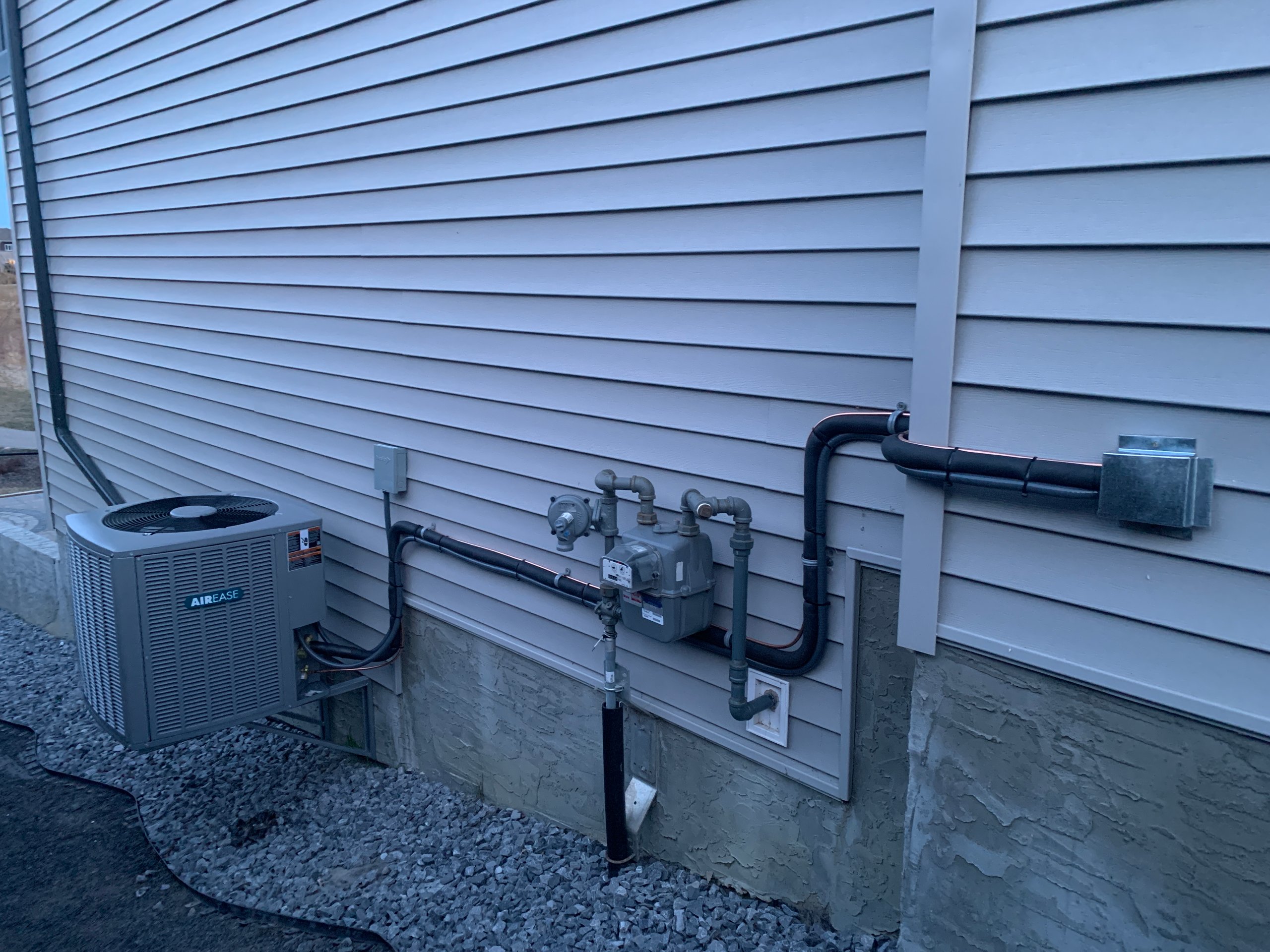 Exterior wall of a house with grey siding featuring an Airease air conditioning unit and gas meter connected by insulated pipes.
