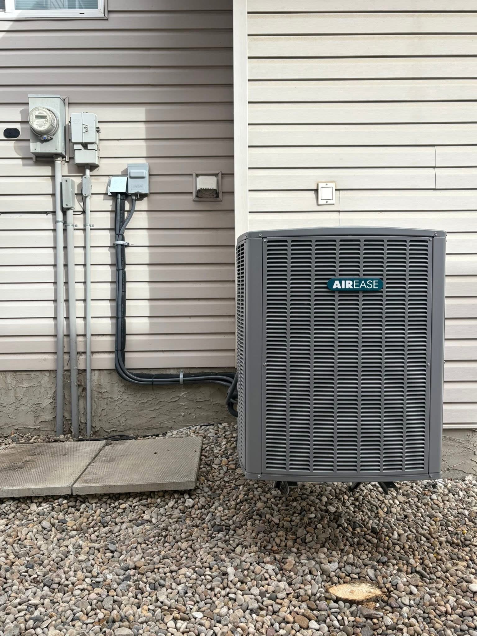 Outdoor air conditioning unit labeled AIR EASE installed against a beige vinyl siding house wall on gravel ground.