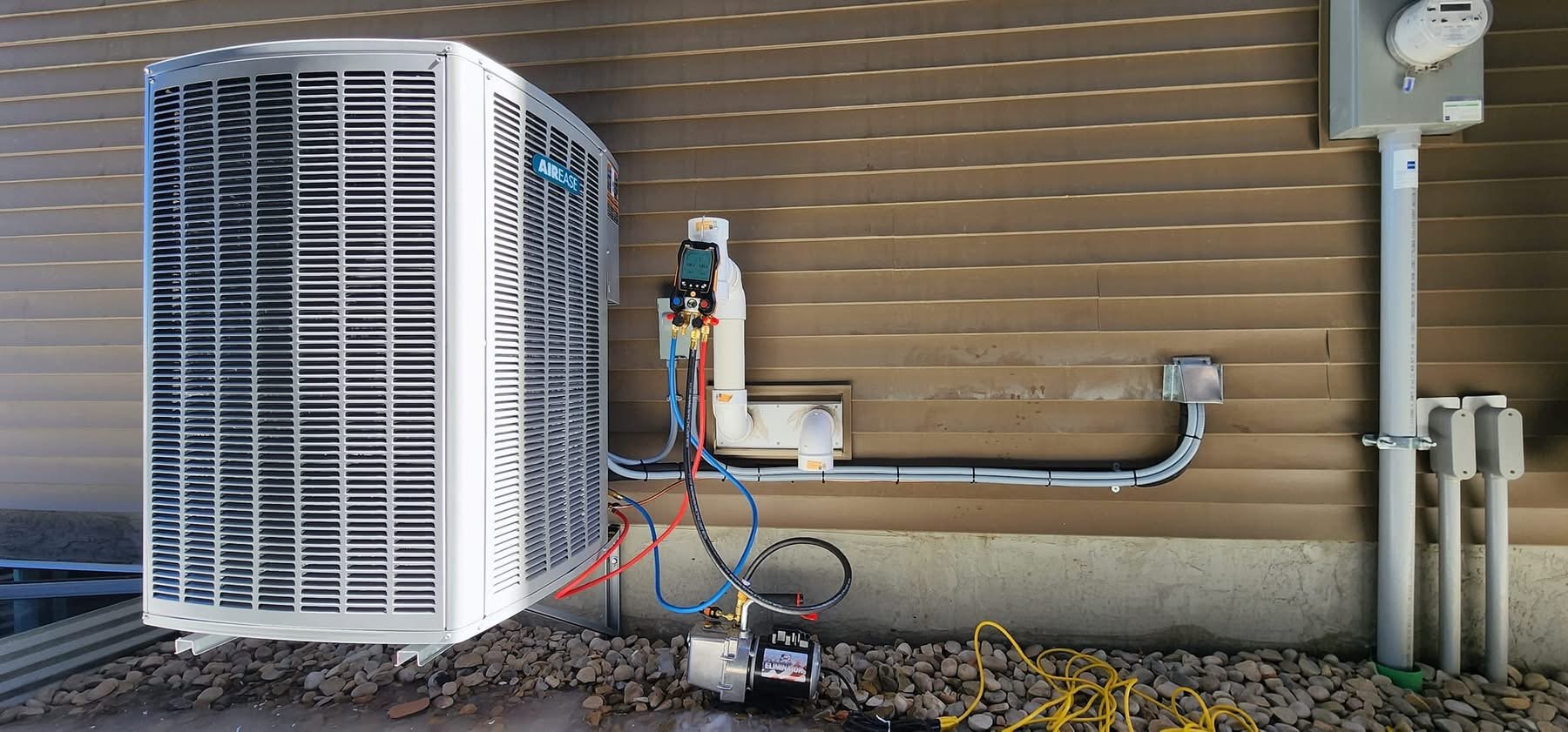 Outdoor air conditioning unit with connected HVAC gauges and compressor pump on rocky ground against a brown siding wall.