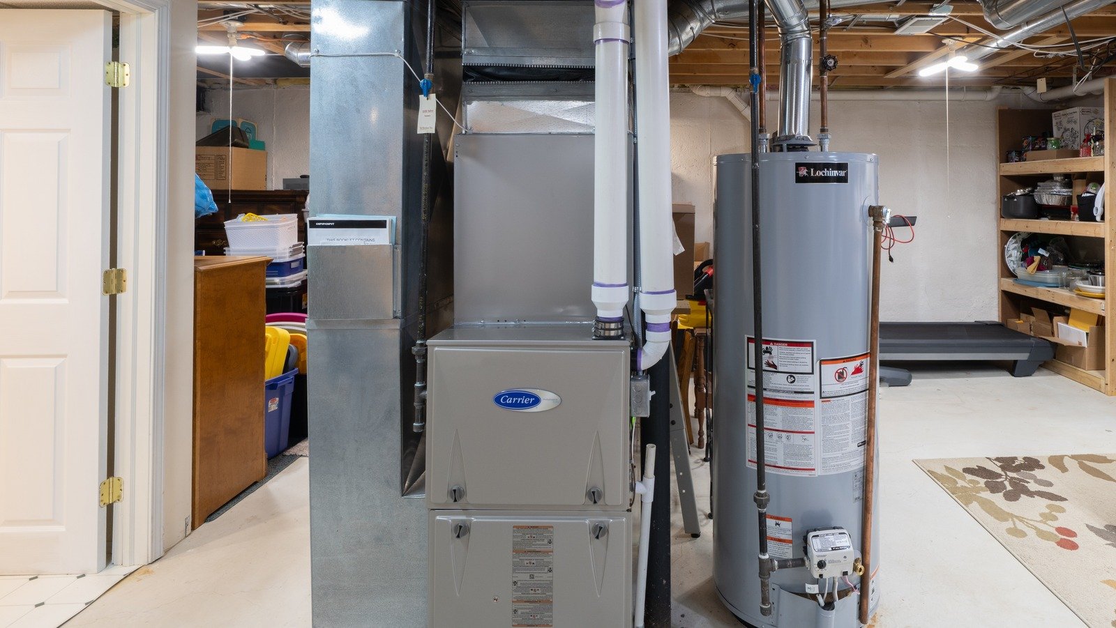 Basement utility room with a Carrier furnace, a Lochinvar water heater, storage shelves with kitchenware, and a treadmill in the background.