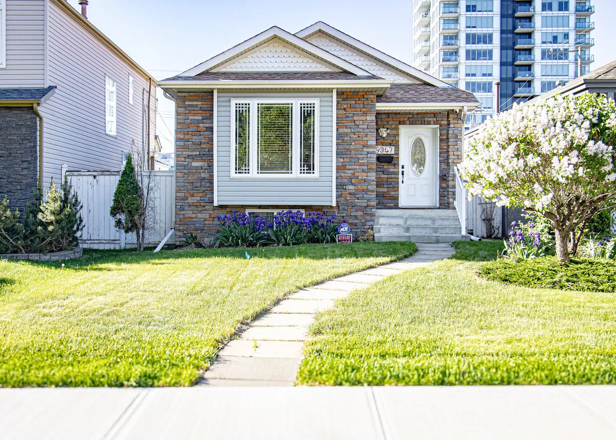 Single-story house with stone and siding exterior, a front garden with purple flowers, trimmed grass, and a white door with an oval window.