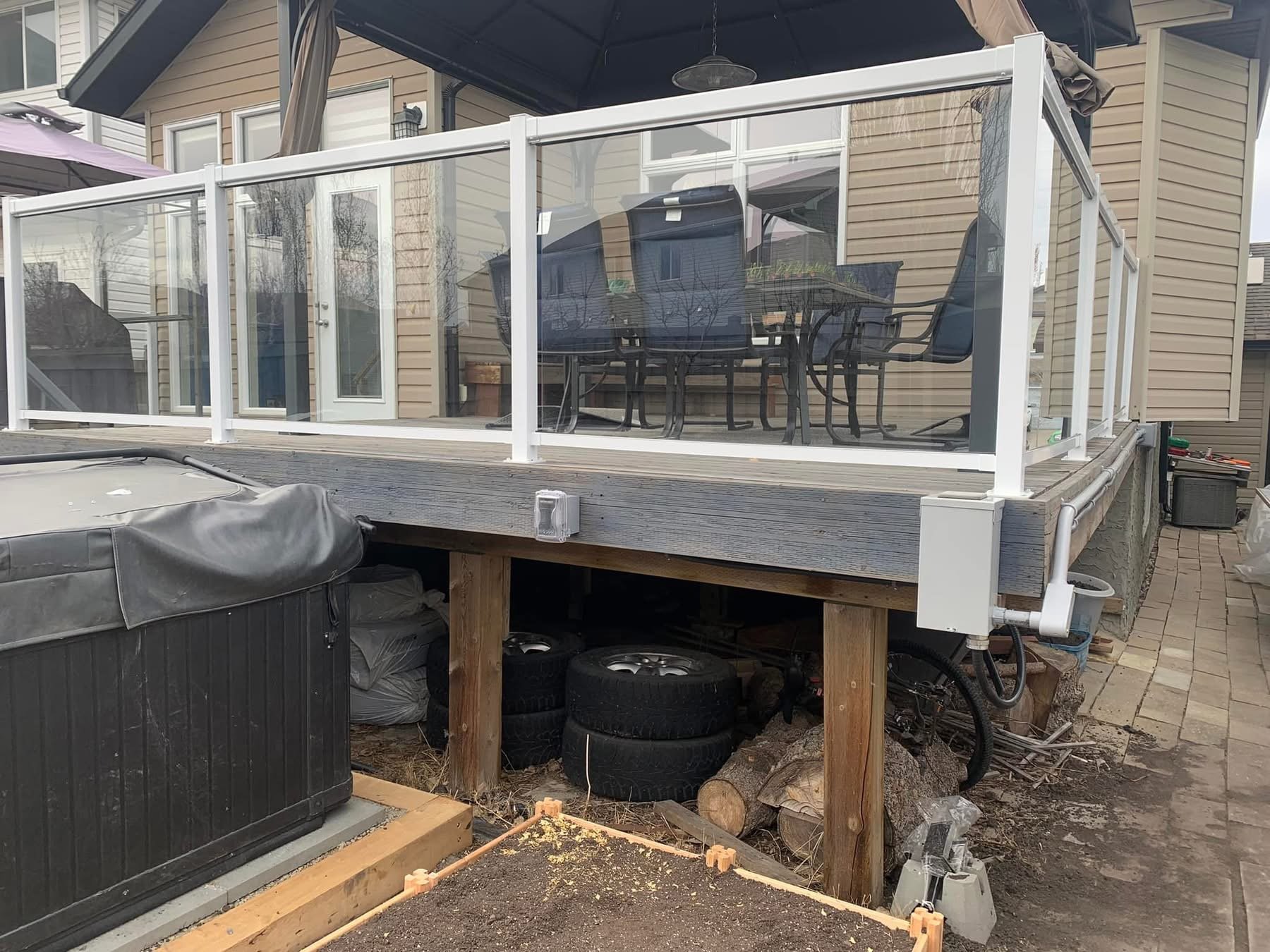 Outdoor patio with glass railing, table and chairs on deck, and stacked tires underneath with a covered hot tub nearby.