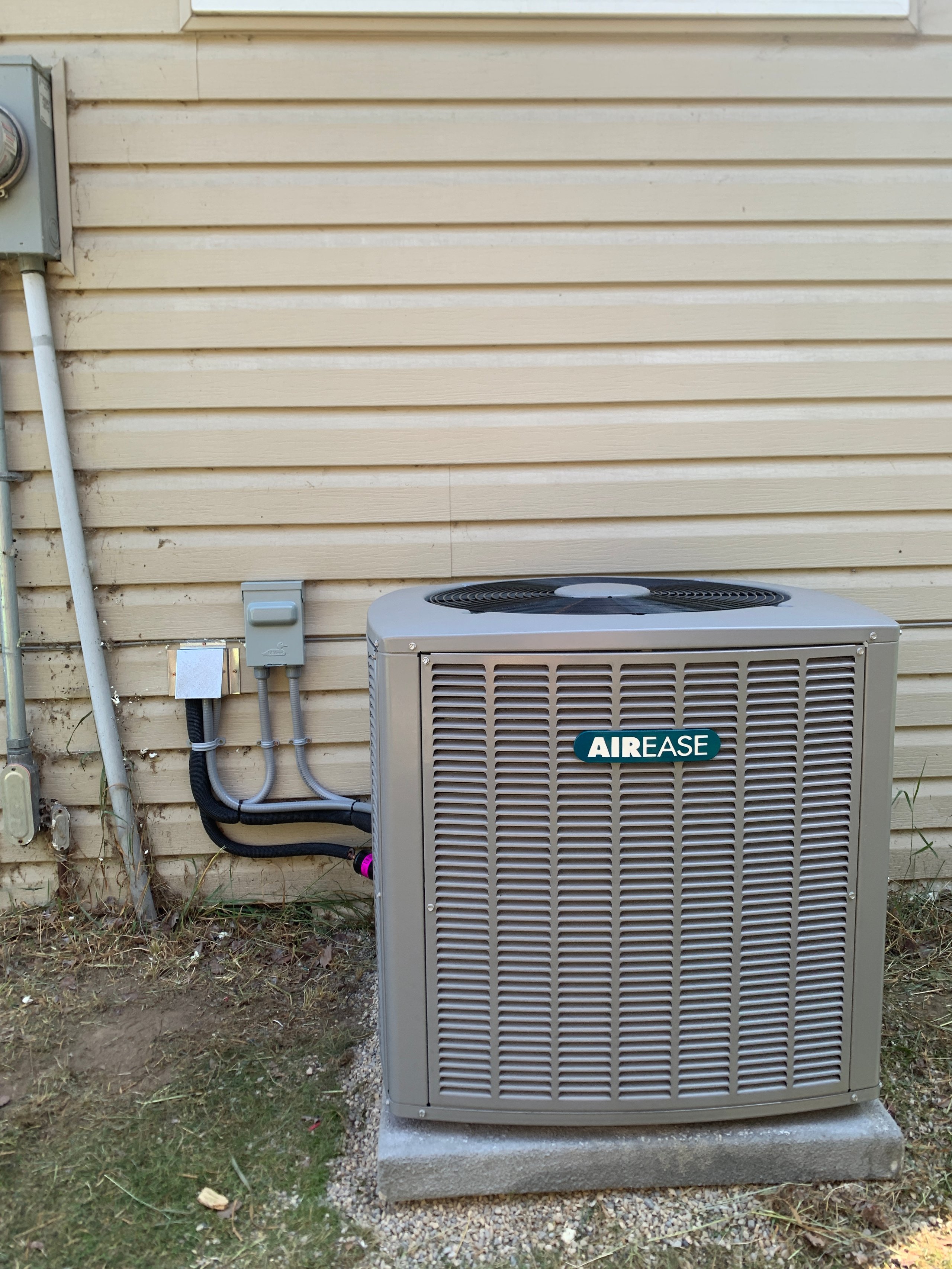 Outdoor AIR EASE air conditioning unit installed on a concrete slab against a beige house wall with connected electrical conduits.