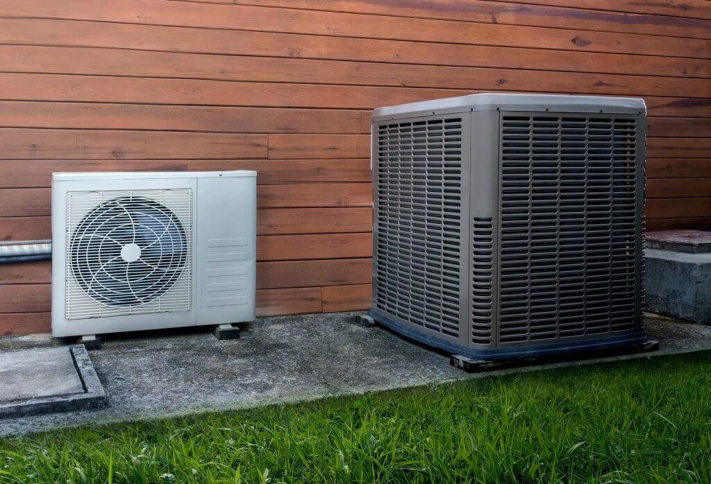 Two outdoor air conditioning units on concrete beside a wooden-paneled house wall and green grass.